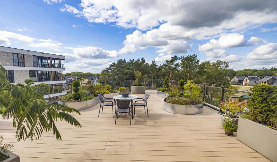 Je vous propose cette balise alt pour l'image : "Vaste terrasse sur toit en bois composite beige clair avec table et chaises métalliques, jardinières circulaires plantées, vue panoramique sur un quartier résidentiel avec immeubles modernes et espaces verts, ciel bleu parsemé de nuages blancs