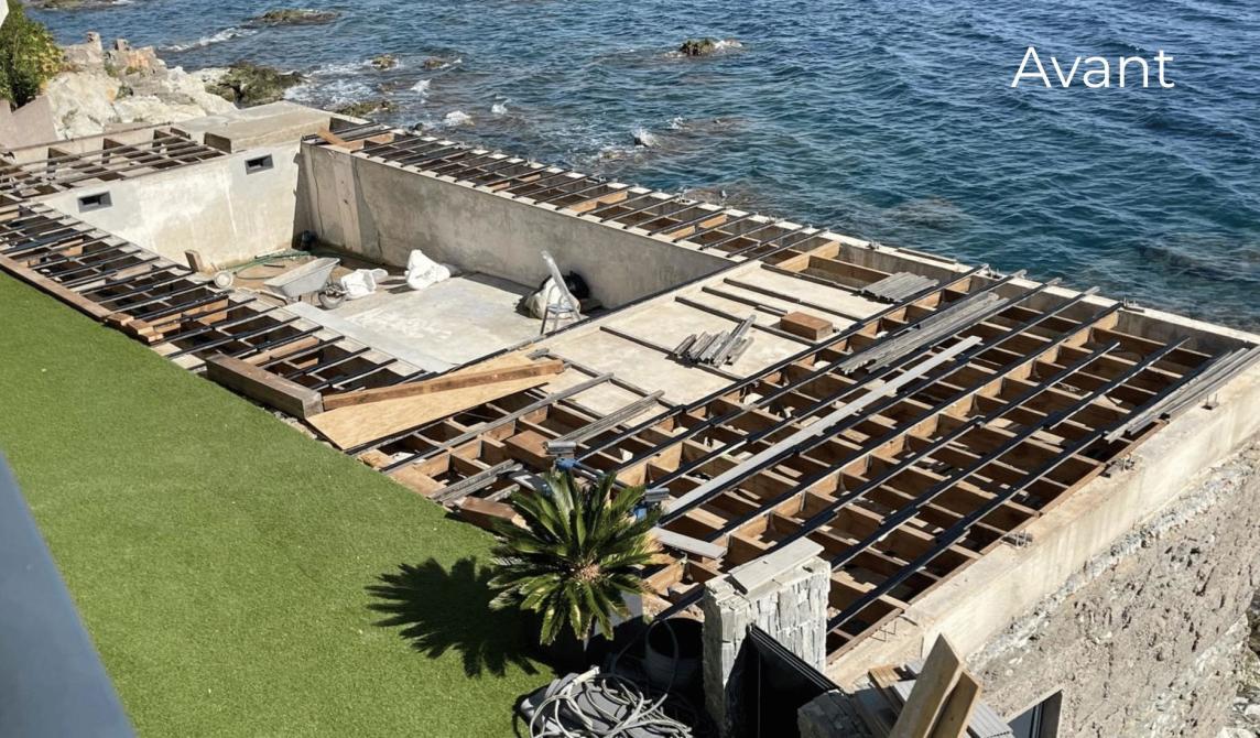 Structure en cours de construction 'Avant' l'installation, montrant l'ossature en bois d'une terrasse en bord de mer avec vue panoramique sur l'eau. La préparation du support pour recevoir les lambourdes en aluminium réversible Réversil de Silvadec. Un palmier est visible au premier plan près de la fondation en béton.