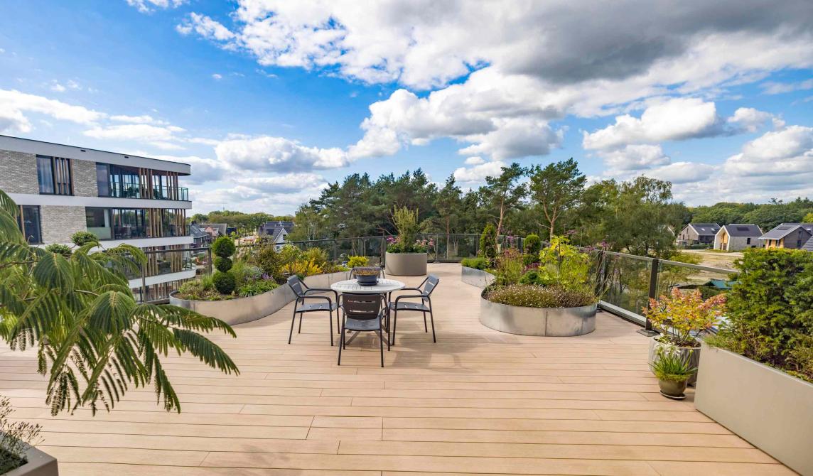 Vaste terrasse sur toit en bois composite beige clair avec table et chaises métalliques, jardinières circulaires plantées, vue panoramique sur un quartier résidentiel avec immeubles modernes et espaces verts, ciel bleu parsemé de nuages blancs